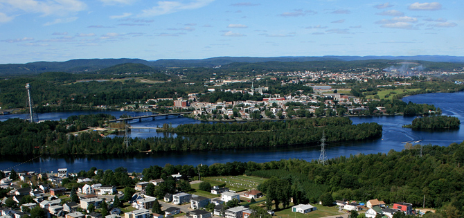 Vue aérienne d'une ville le long d'une rivière entourée de collines verdoyantes, ciel bleu avec nuages épars